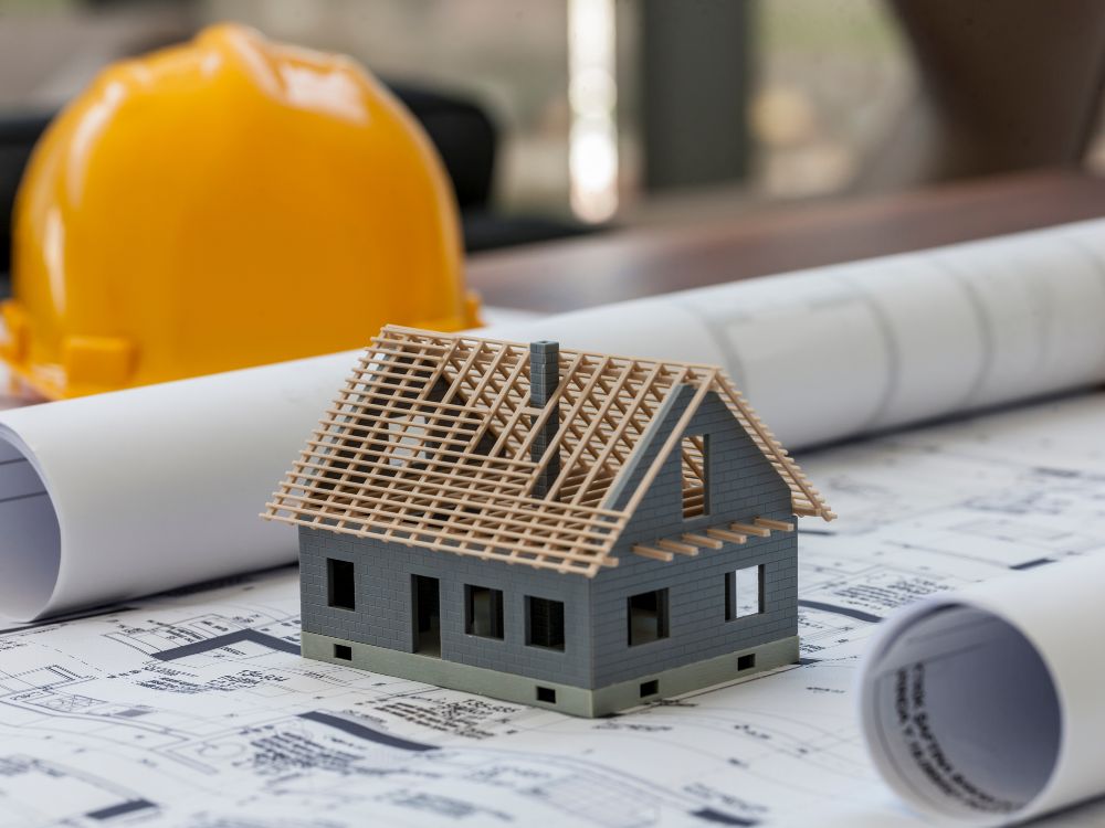 Maquete de casa sobre planta arquitetônica com capacete de obra ao fundo, representando projeto e construção de imóveis residenciais.